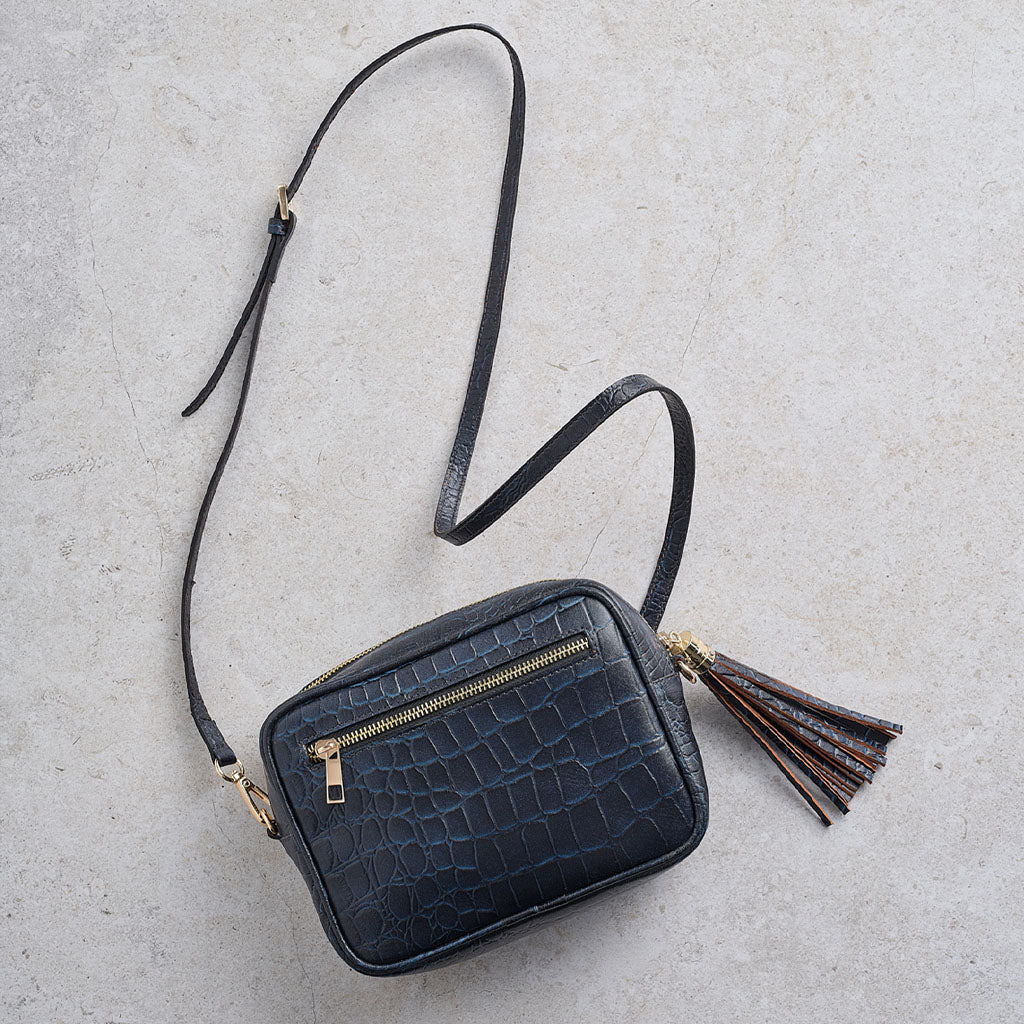 A small black crocodile-embossed leather crossbody bag with a gold zipper, a long black shoulder strap, and a brown and black tassel charm, laid flat on a light gray textured background.