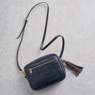 A small black crocodile-embossed leather crossbody bag with a gold zipper, a long black shoulder strap, and a brown and black tassel charm, laid flat on a light gray textured background.
