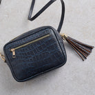 A dark blue crocodile-embossed leather crossbody bag with gold-tone hardware, a top zip closure, and a decorative tassel accent, shown on a light gray textured background.