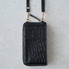 Black crocodile-embossed leather zip-around phone wallet with a matching shoulder strap and gold-tone hardware, shown flat on a light textured surface.