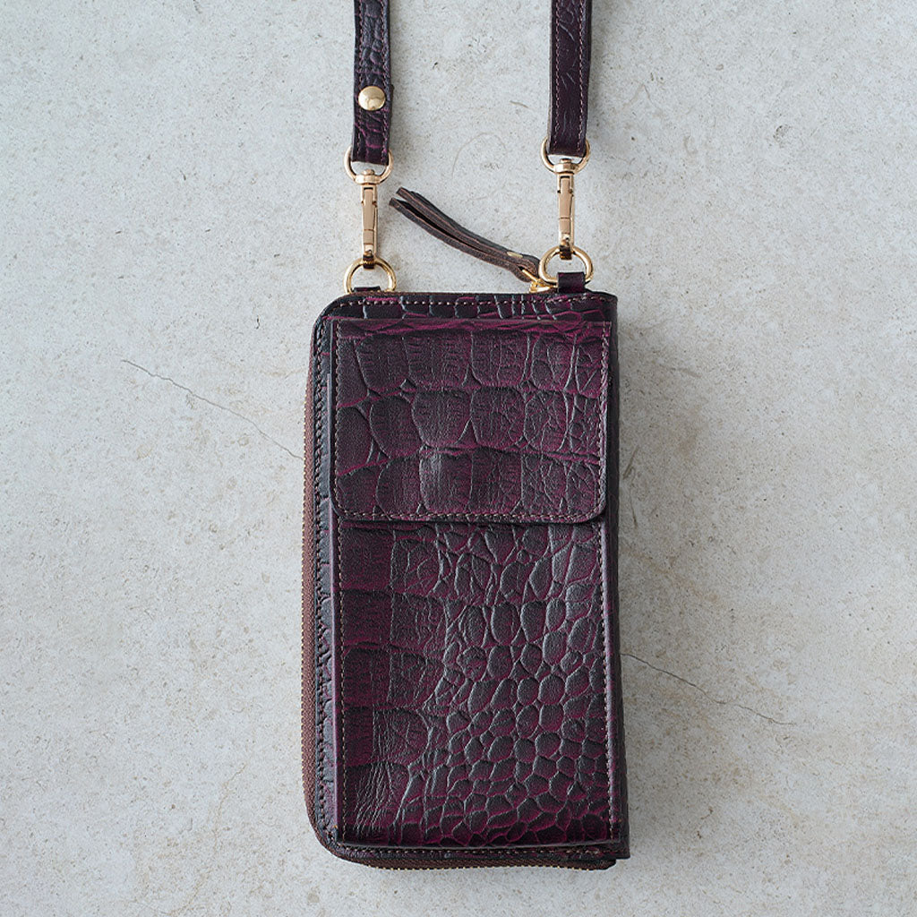 A dark purple crocodile-embossed leather phone pouch with a matching shoulder strap and gold-tone hardware, laid flat on a light gray textured surface.