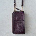A dark purple crocodile-embossed leather phone pouch with a matching shoulder strap and gold-tone hardware, laid flat on a light gray textured surface.