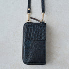A black crocodile-embossed leather zip-around phone wallet with a matching wrist strap and gold-tone hardware, laid flat on a light textured surface.