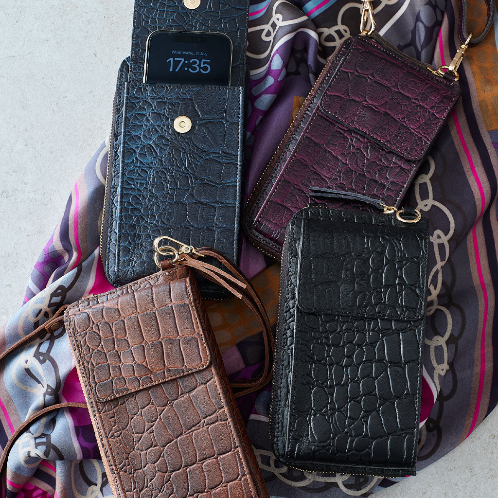 Three embossed leather wallet phone cases in brown, black, and deep purple, arranged on a patterned silk scarf with gold-tone hardware.