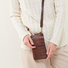A person wearing a cream cable-knit sweater and beige pants holds a brown crocodile-embossed leather crossbody phone bag with a matching strap against a light beige background.