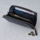 A black embossed leather zip-around wallet shown open, revealing multiple interior compartments, card slots, and a gold-tone zipper. Euro banknotes and coins are placed inside and beside the wallet on a light stone surface.
