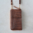 A brown crocodile-embossed leather phone wallet with a matching shoulder strap and gold-tone hardware, laid flat on a light gray textured surface.