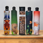 Five folded umbrellas with black handles are lined up on a wooden shelf against a light gray wall. Each umbrella cover has a different printed design: a colorful abstract floral pattern, a green background with a highland cow portrait, a leopard print with a small bird, a dark green and orange leaf pattern, and a pink-to-orange gradient with a bird.