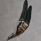 A decorative feather pin featuring two long iridescent feathers with black, brown, and cream bands, attached to an ornate antique bronze safety pin, displayed on a textured gray background.