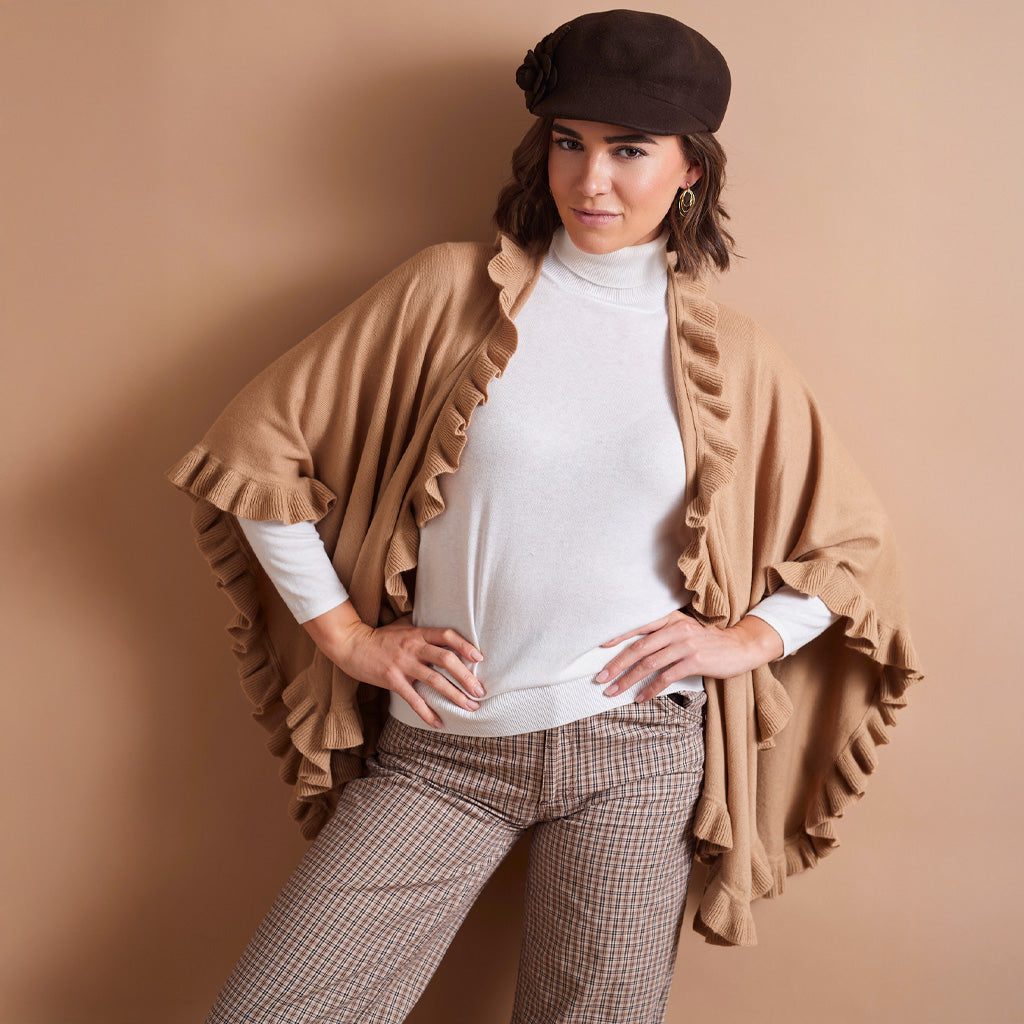 A woman models a tan ruffled cape-style cardigan over a white turtleneck, paired with brown check trousers and a dark brown beret, against a solid tan studio background.