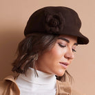 A woman wearing a dark brown felt-style newsboy cap with a large fabric rose embellishment on the side, paired with a white turtleneck and tan jacket against a plain beige background.