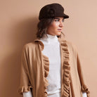A woman wearing a brown felt newsboy cap with a fabric flower, a white ribbed turtleneck sweater, and a camel beige open-front cardigan with ruffle trim, posed against a solid warm beige background.