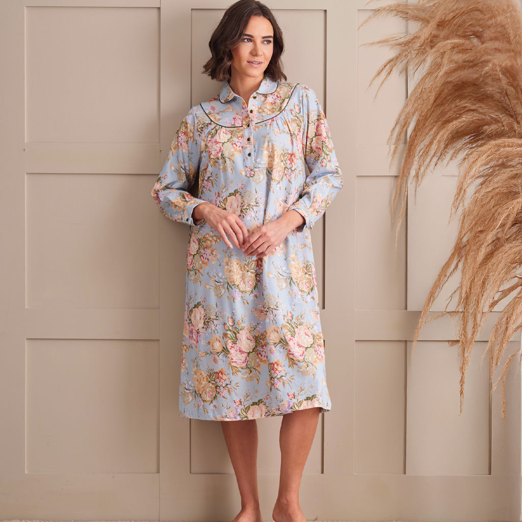 A woman wearing a light blue long-sleeve nightgown with a pink and beige floral print, standing against a beige paneled wall with dried pampas grass on the right.