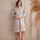 A woman wearing a light blue long-sleeve nightgown with a pink and beige floral print, standing against a beige paneled wall with dried pampas grass on the right.