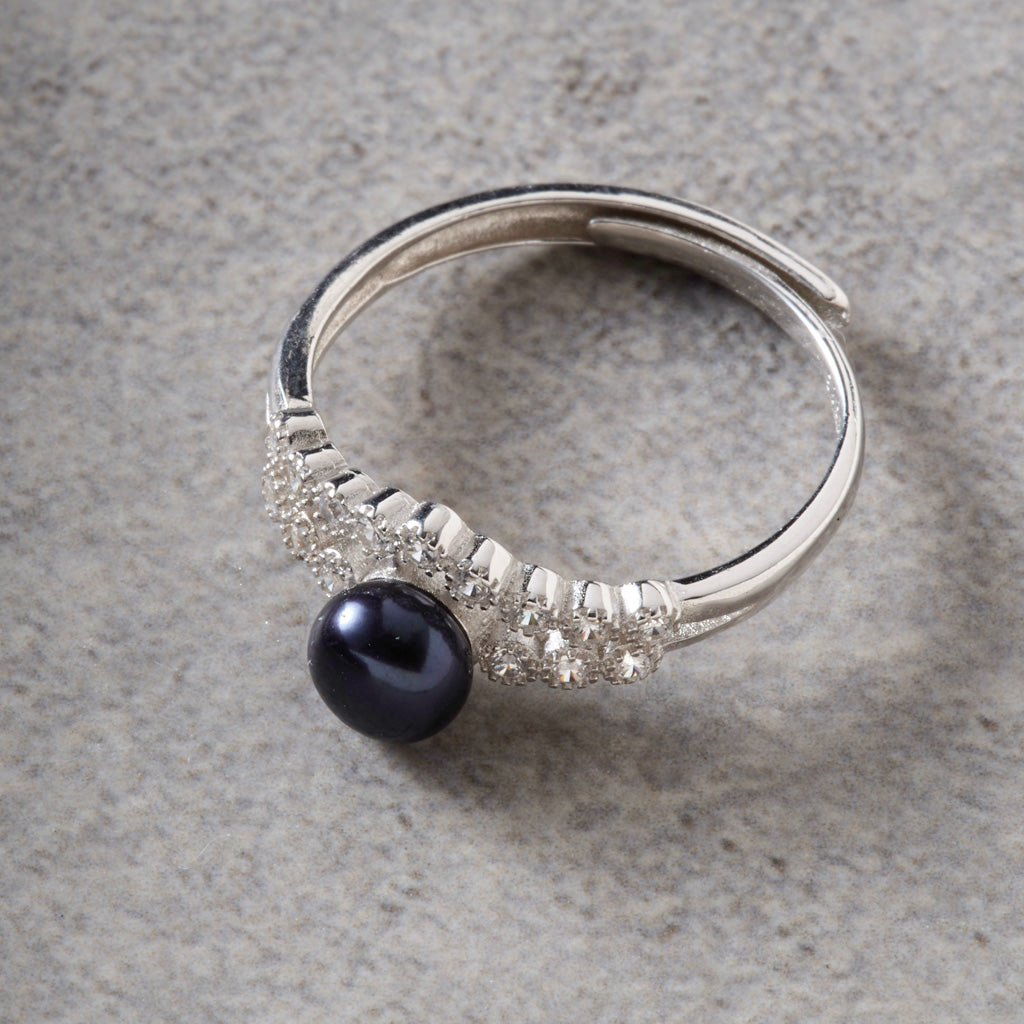A silver-toned open band ring featuring a curved row of small clear stones and a single dark black pearl at the center, photographed on a textured gray surface.