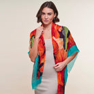 A woman modeling a lightweight, multicolored abstract print shawl with fringed edges over a light gray dress, set against a plain beige studio background.