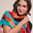 A woman wearing a lightweight, multicolored abstract print scarf with bright turquoise, orange, red, and black tones, photographed against a solid light beige background.