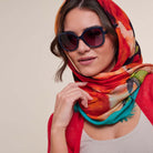 A woman wearing a vibrant, multicolored abstract print scarf in shades of red, orange, teal, and green, styled with dark sunglasses and a red cardigan against a neutral beige background.