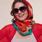 A smiling woman wearing a vibrant multicolor abstract print scarf in shades of orange, red, teal, and black, styled with dark sunglasses and a red open-front cardigan.