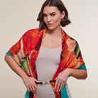 A woman modeling a vibrant, multicolored abstract print shawl draped over her shoulders, worn over a light beige dress. The shawl features a mix of red, orange, yellow, green, and teal colors in a painterly pattern.