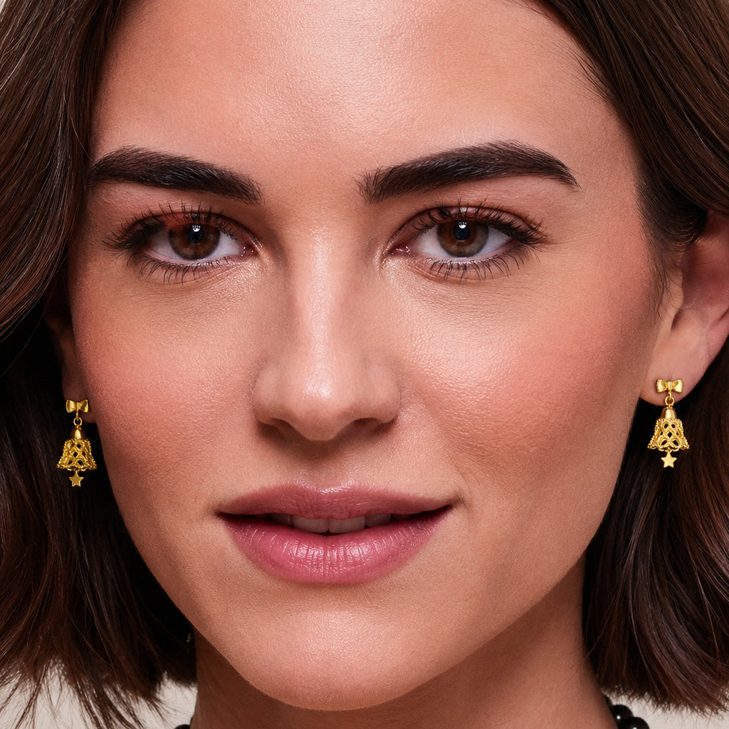 A close-up of a woman wearing a pair of gold dangle earrings with a bell and star design, shown against her face.