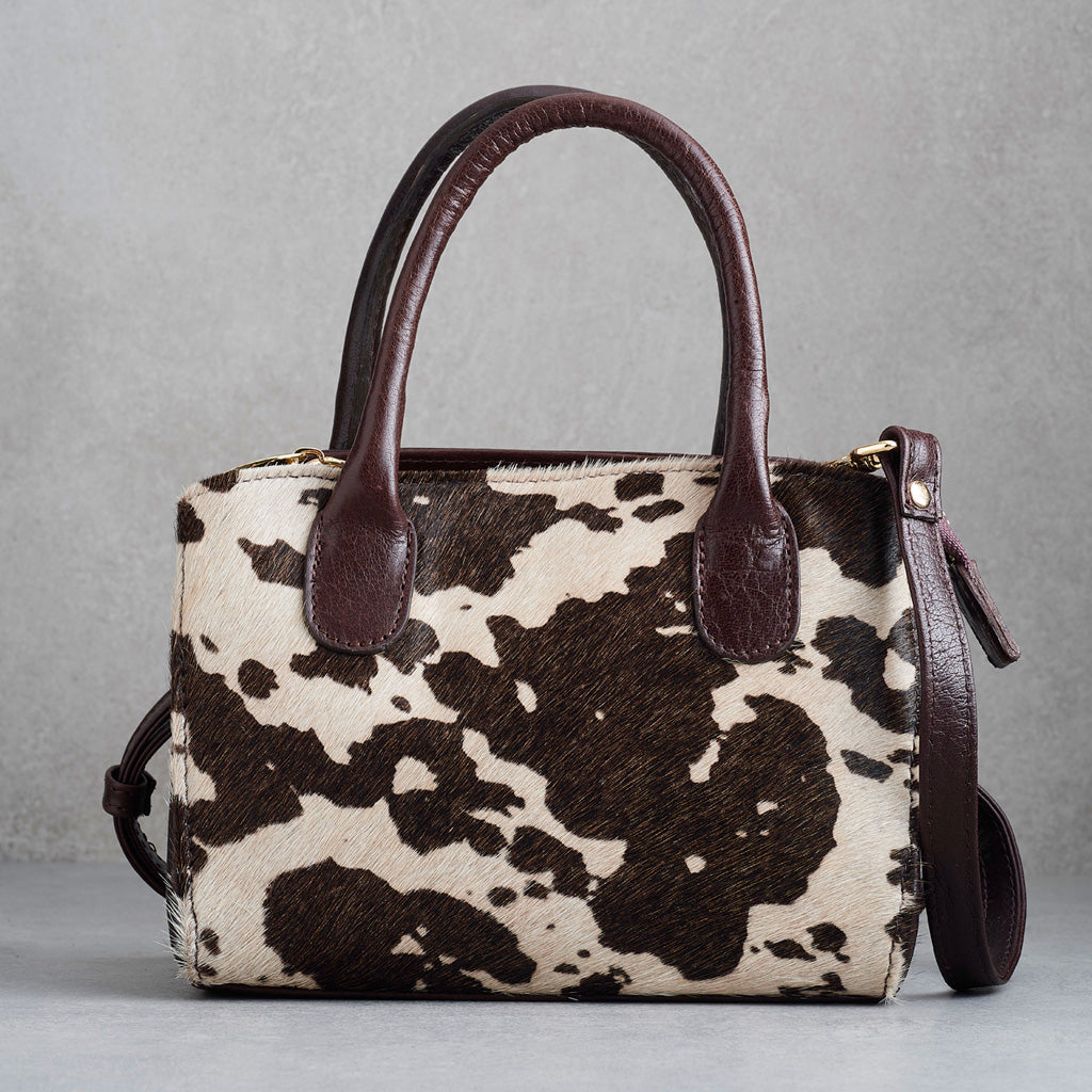 A structured handbag with a brown and cream cowhide pattern body and dark brown leather handles, shown against a neutral gray background.