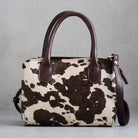 A structured handbag with a brown and cream cowhide pattern body and dark brown leather handles, shown against a neutral gray background.