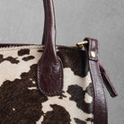 Close-up of a cowhide handbag with dark brown leather handles and trim, featuring a brown, black, and cream cow print pattern and gold-tone hardware.
