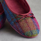 A close-up of a pair of knit slip-on flats with a multicolor plaid pattern in blue, pink, yellow, and green, featuring a dusty rose pink suede-like finish and matching bow details.