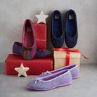 Four pairs of slip-on flat shoes arranged with gift boxes and star decorations against a neutral gray background. The shoes include multicolor plaid, dark navy blue, and lavender purple herringbone patterns, each with a matching fabric bow on the toe.