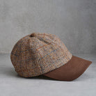 A classic six-panel tweed baseball cap with a brown leather visor and button, shown from a side profile on a gray textured background.