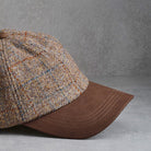 A close-up of a baseball cap featuring a textured tweed crown in beige, brown, and muted blue with a solid brown curved brim, set against a gray concrete background.