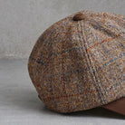 A close-up of a brown tweed flat cap with a brown button on top, resting on a gray surface against a gray wall.