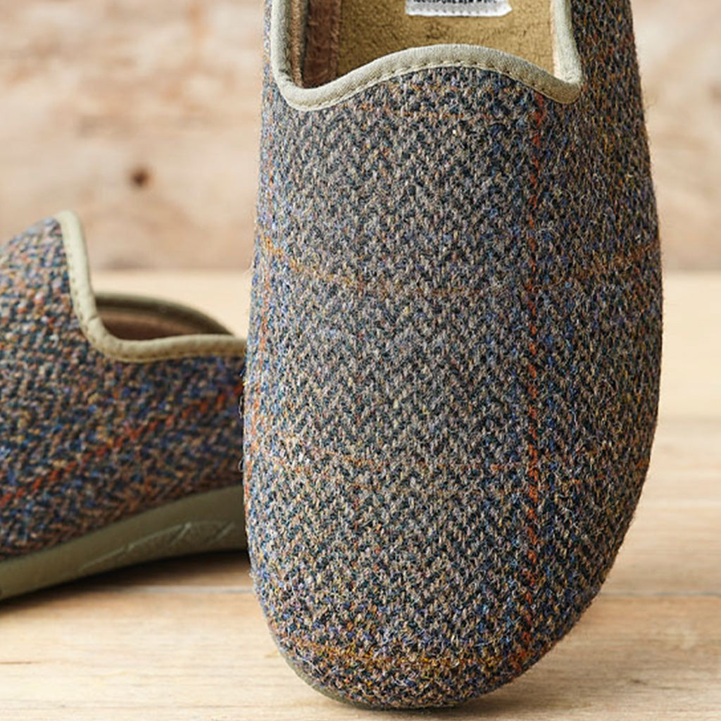 A close-up of a slip-on house shoe in textured tweed fabric with a blue, gray, and brown herringbone pattern, shown on a light wood surface with another shoe blurred in the background.