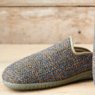 A single slip-on mule shoe made from a textured herringbone tweed fabric in muted blue, gray, and brown tones, with an olive green sole, shown on a light wood surface.