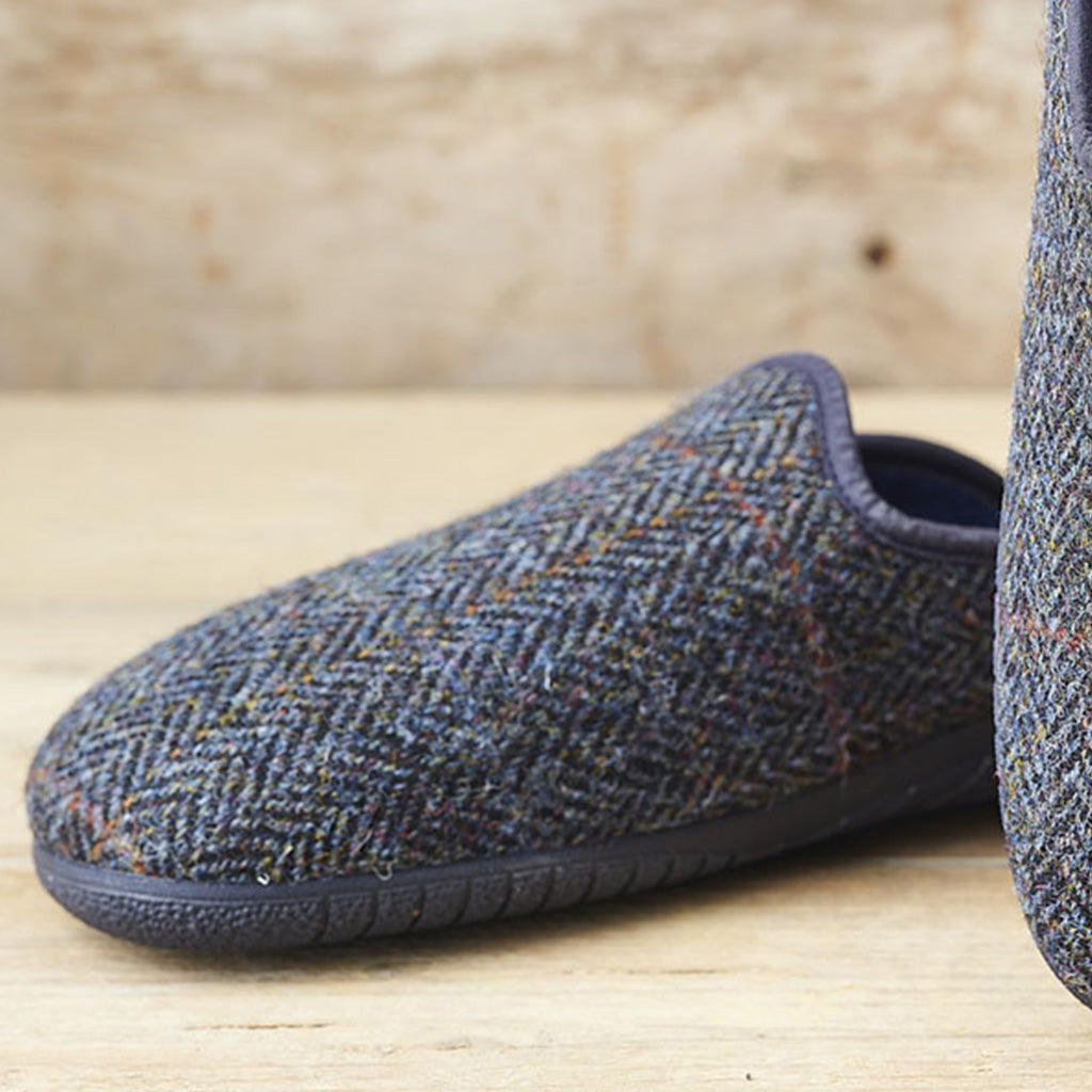 A pair of slip-on slippers in a dark blue-gray herringbone tweed fabric, shown on a light wood surface with a rustic wooden background.