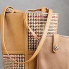 A close-up of a structured tote bag with tan leather trim and handles, featuring a woven houndstooth pattern panel in beige, brown, and muted red. A matching tan leather snap-button strap or pouch is partially visible in the foreground against a soft gray background.