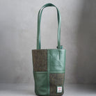 A green leather and herringbone tweed bucket bag with dual shoulder straps and a visible Woolworths label patch on the lower front, photographed against a neutral gray background.