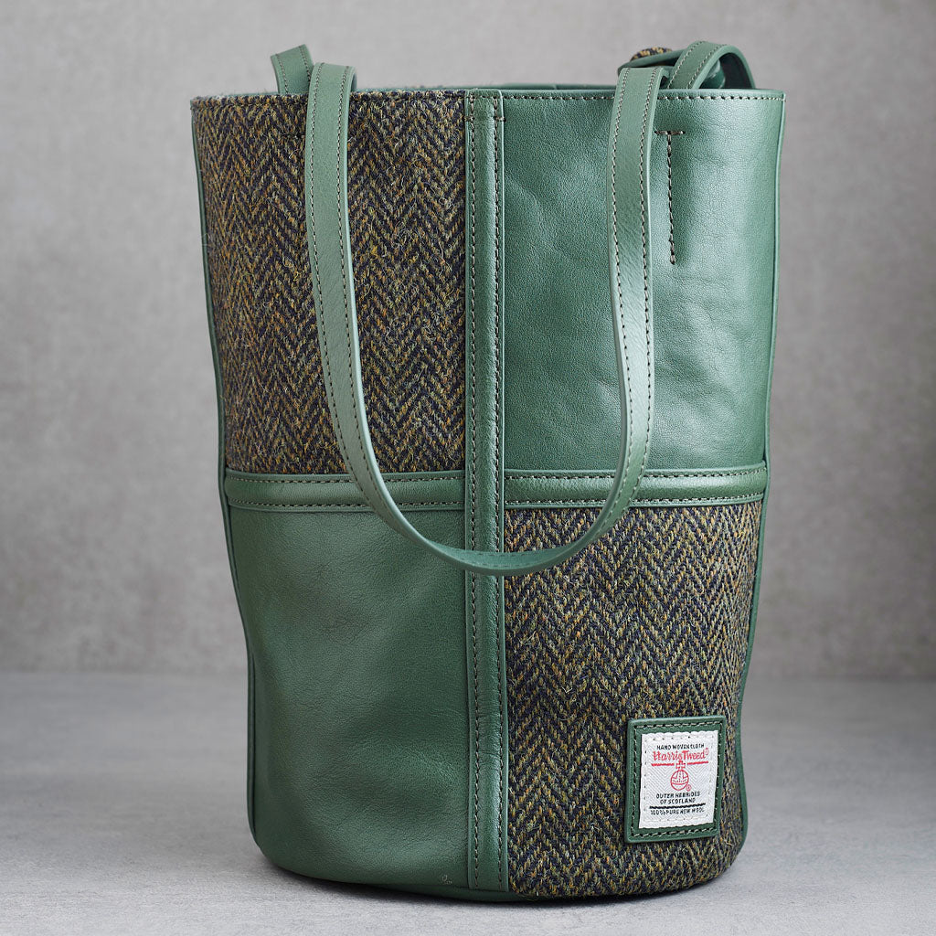 A green leather and herringbone tweed bucket bag with dual shoulder straps and a visible Woolworths label patch on the lower front, photographed against a neutral gray background.