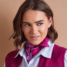 A woman with brown hair wearing a bright magenta and purple patterned neck scarf, a white and blue striped button-down shirt, and a textured magenta knit vest, set against a solid warm beige background.