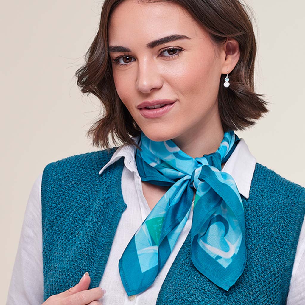 A woman wearing a teal blue patterned silk scarf tied around her neck, styled over a white collared shirt and a textured teal knit vest, against a solid light beige background.