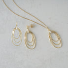 Gold-toned jewelry set featuring a delicate chain necklace and matching drop earrings, each with three nested oval loops, displayed on a light gray textured surface.