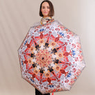 A woman holding up a large open umbrella to display its intricate pattern. The umbrella features a symmetrical mandala-style design with red, orange, black, and blue motifs on a white background.