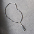 A beaded necklace with small clear faceted beads and a light gray stone pendant, laid on a textured gray surface.