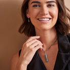 A smiling woman with brown hair models a silver-toned necklace featuring a gray stone pendant, wearing a black top against a warm brown background.