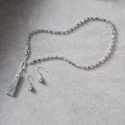 A beaded necklace and matching drop earrings laid out on a textured light gray surface. The jewelry features small clear faceted beads with silver-toned metal findings and a light gray tassel charm.