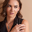 A close-up of a woman wearing a silver-toned chain necklace with a gray stone pendant, along with matching drop earrings, against a warm beige background.