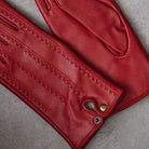 A close-up of a pair of red leather gloves laid flat on a textured gray surface, showing detailed stitching and brass snap closures.