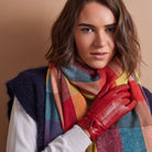 A woman wearing a chunky knit sweater vest, a vibrant multicolor plaid scarf, and a bright red leather glove, set against a solid warm beige background.