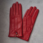 A pair of red leather gloves laid flat on a textured gray background, showing the back of one glove with decorative stitching and a small gold-tone snap closure, and the palm side of the other glove.